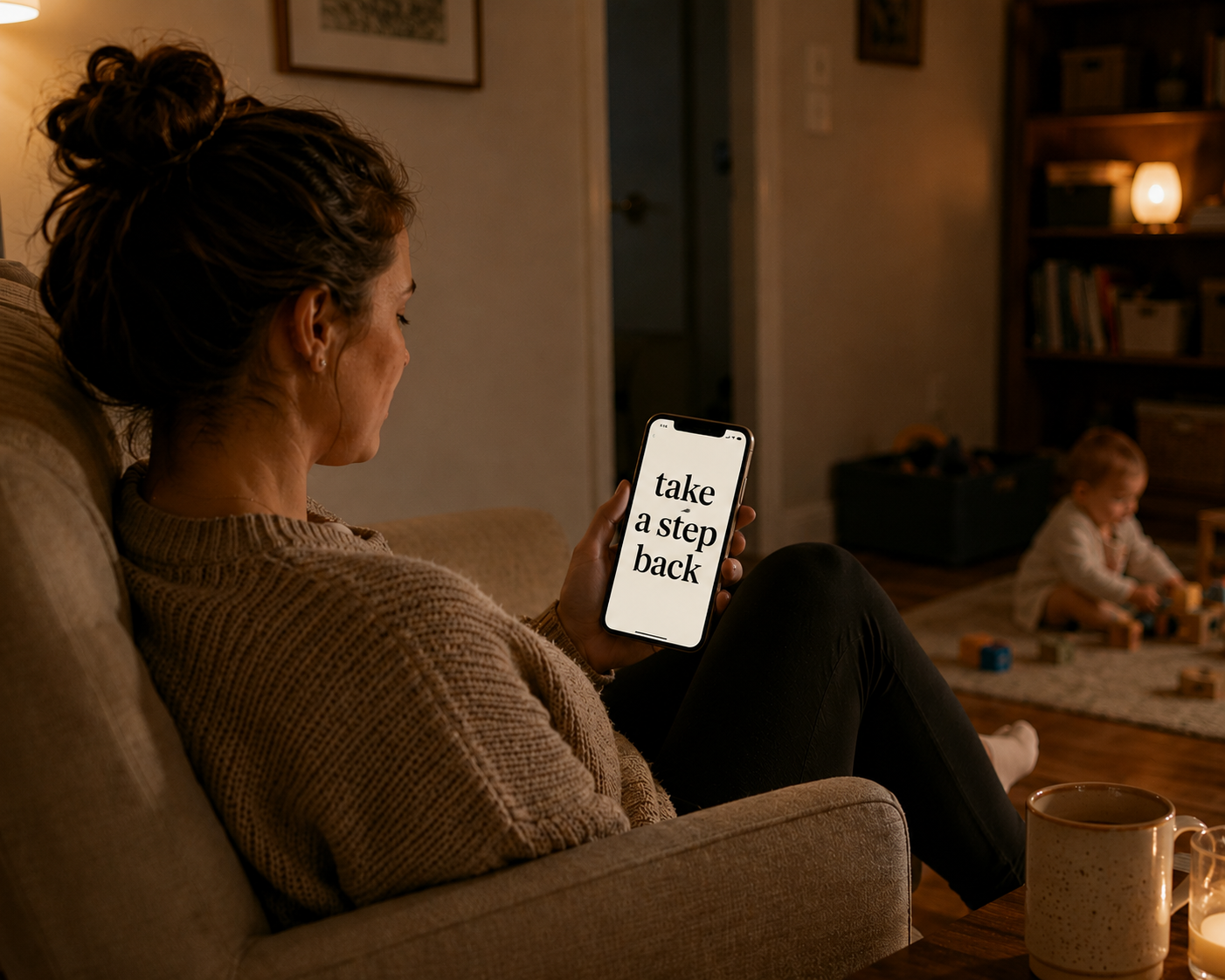 A parent on the couch at night holding a phone that reads 'take a step back' while a baby plays nearby